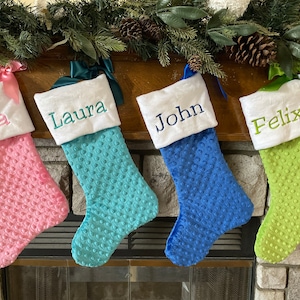 May include: Four personalized Christmas stockings with white felt cuffs and embroidered names: Lila, Laura, John, and Felix. The stockings are pink, teal, blue, and green with a textured, bubble-like fabric.