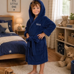 May include: A navy blue hooded bathrobe made of soft, plush fabric. The robe features long sleeves, a tie belt at the waist, and two front pockets. The name Nick is embroidered in white thread on the upper left chest area of the garment.