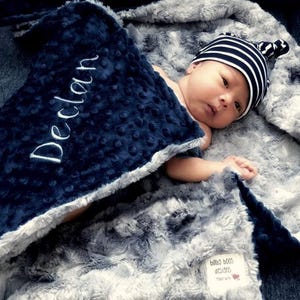 May include: A baby wrapped in a soft, gray and navy blue blanket. The navy blue blanket has the name "Declan" embroidered in white. The baby wears a striped beanie. The blanket is made by "baby boos designs".