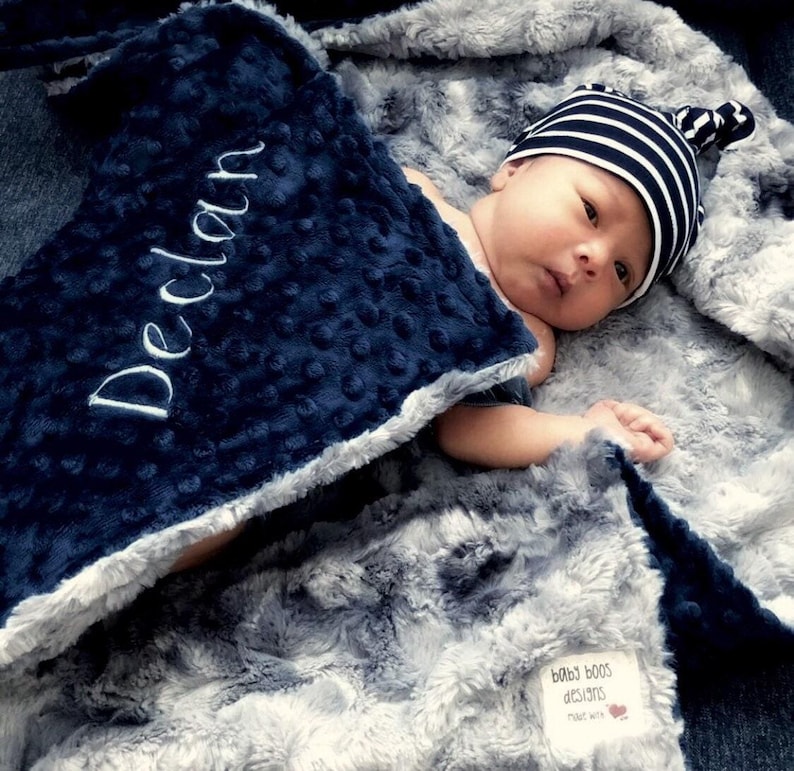 May include: A baby wrapped in a soft, gray and navy blue blanket. The navy blue blanket has the name "Declan" embroidered in white. The baby wears a striped beanie. The blanket is made by "baby boos designs".
