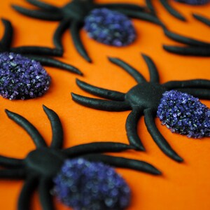 May include: Black spider decorations with purple sugar crystal bodies on an orange background. The spiders have eight legs and are arranged in a scattered pattern, ideal for Halloween or themed parties.