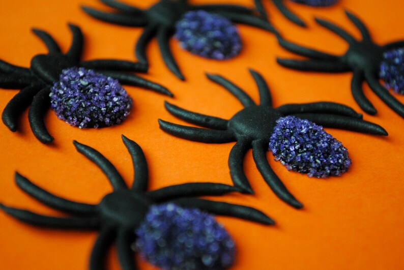 May include: Black spider decorations with purple sugar crystal bodies on an orange background. The spiders have eight legs and are arranged in a scattered pattern, ideal for Halloween or themed parties.