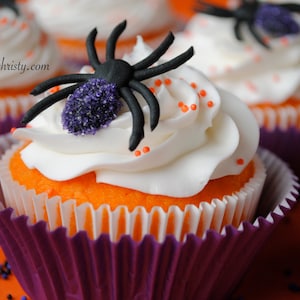 May include: Halloween cupcakes with orange sponge, white icing, and a black spider decoration with purple detailing. The cupcakes are in purple and white paper cases, with orange sprinkles.