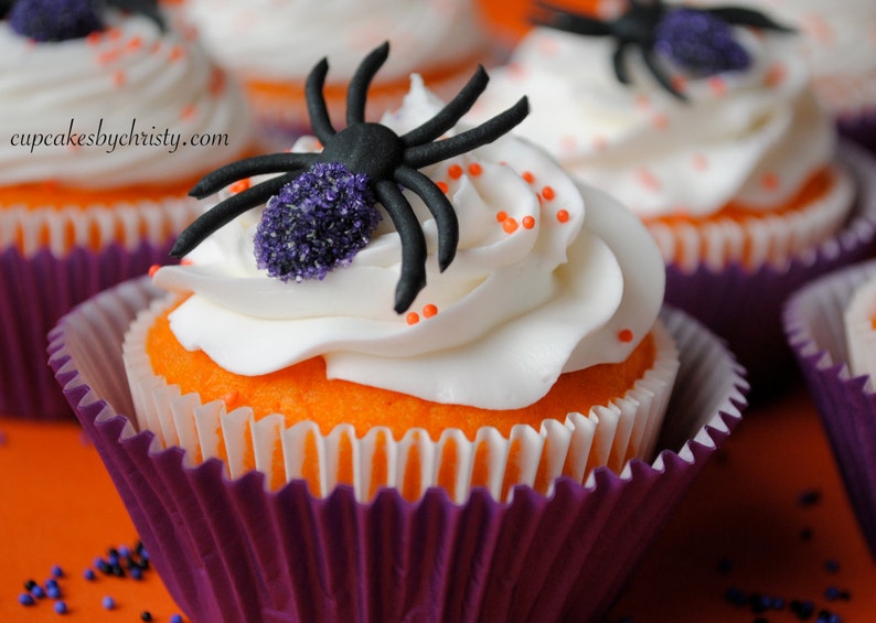 May include: Halloween cupcakes with orange sponge, white icing, and a black spider decoration with purple detailing. The cupcakes are in purple and white paper cases, with orange sprinkles.