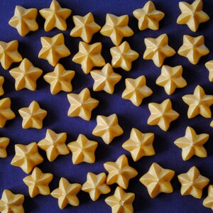 Puede incluir: Una colecci&oacute;n de peque&ntilde;os objetos en forma de estrella de color amarillo c&aacute;lido, dispersos sobre una superficie azul oscuro. Las estrellas tienen un aspecto texturizado y tridimensional, creando un patr&oacute;n visualmente atractivo. La imagen est&aacute; bien iluminada, destacando los detalles de cada estrella.