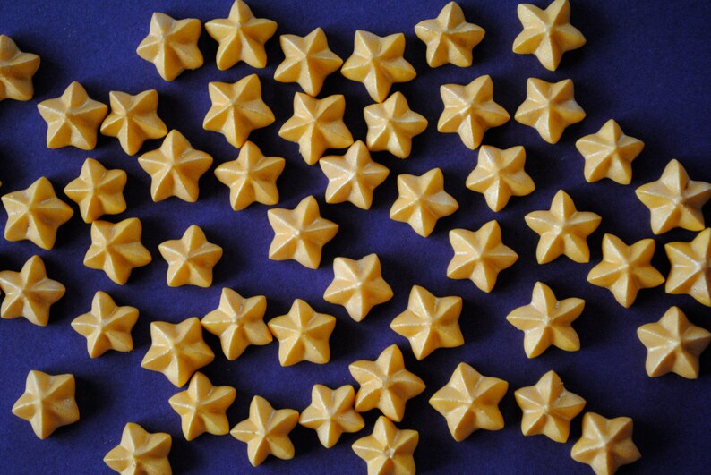 Puede incluir: Una colecci&oacute;n de peque&ntilde;os objetos en forma de estrella de color amarillo c&aacute;lido, dispersos sobre una superficie azul oscuro. Las estrellas tienen un aspecto texturizado y tridimensional, creando un patr&oacute;n visualmente atractivo. La imagen est&aacute; bien iluminada, destacando los detalles de cada estrella.