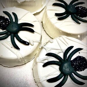 May include: Close-up of four white frosted treats decorated for Halloween. Each treat has a black spider and a web design made with white icing. A cluster of black sprinkles adds detail to each treat.