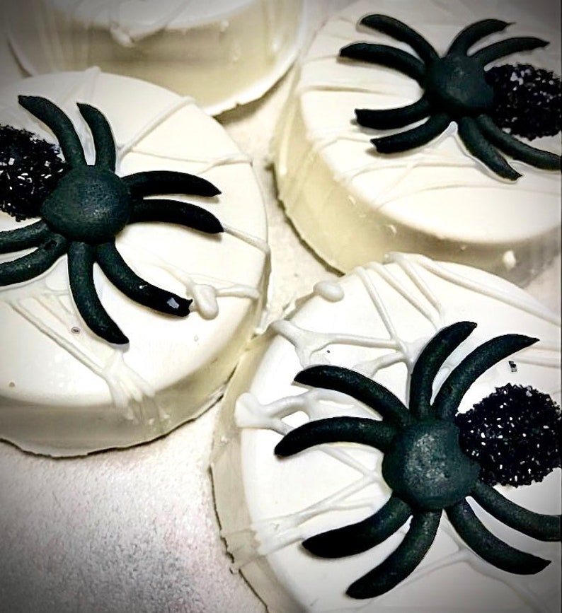May include: Close-up of four white frosted treats decorated for Halloween. Each treat has a black spider and a web design made with white icing. A cluster of black sprinkles adds detail to each treat.