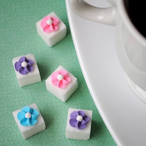 May include: Close-up of white sugar cubes decorated with colorful flower designs. The flowers are pink, purple, and blue with white centers. A white coffee cup and saucer are in the background, set against a light green surface.