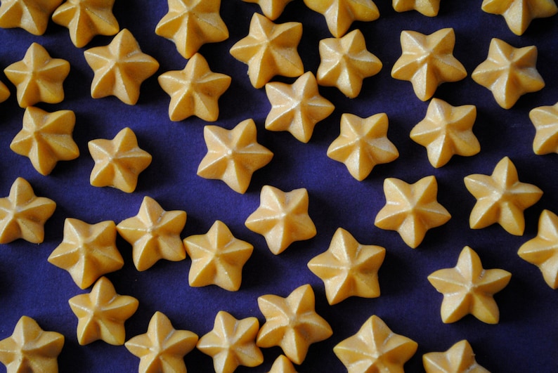Puede incluir: Primer plano de numerosos caramelos en forma de estrella de color amarillo dorado. Los caramelos est&aacute;n dispuestos sobre una superficie azul oscuro, creando un contraste atractivo. Estas golosinas son probablemente para hornear o decorar.