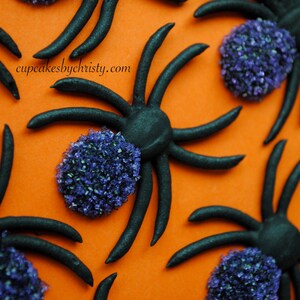 May include: An orange surface decorated with black spiders and purple, granular accents. The spiders have eight legs and are arranged in a repeating pattern. The text "cupcakesbychristy.com" is visible in the image.