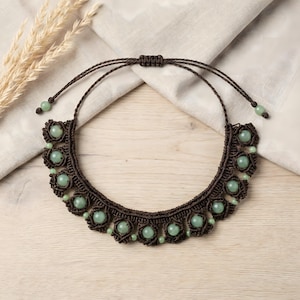 May include: A dark brown macrame necklace featuring a series of round, pale green beads integrated into the knotted cord design. The necklace includes an adjustable sliding knot closure with two thin cords ending in small green beads at the tips.