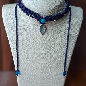 May include: A dark blue macrame choker necklace with a turquoise bead and a silver leaf pendant. The necklace has a braided design and is displayed on a mannequin. The necklace has two long, dangling cords with blue beads at the ends.