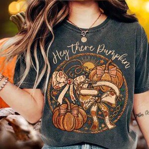 Puede incluir: Una camiseta gris carbón con un gráfico circular y el texto "Hey there Pumpkin". El diseño incluye dos calabazas naranjas, lazos decorativos con patrones florales, un borde de cuerda y pequeños detalles de estrellas en tonos cálidos.