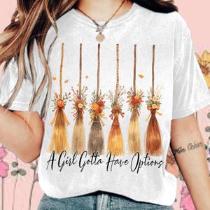 Op de afbeelding: Wit T-shirt met zes decoratieve bezems versierd met bloemstukken en linten. De bezems zijn in bruin-, oranje- en geeltinten. De tekst "A Girl Gotta Have Options" staat eronder.