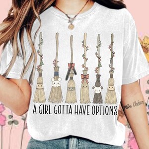 May include: White t-shirt featuring a whimsical design of six cartoon brooms with smiling faces and decorative accents. The brooms are arranged vertically, each with a unique look. The text "A GIRL GOTTA HAVE OPTIONS" is printed below the brooms.
