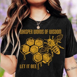 May include: Black t-shirt with a yellow honeycomb and bee graphic. The text "WHISPER WORDS OF WISDOM" is above the honeycomb, and "LET IT BEE" is below. The design features a detailed illustration of a bee.