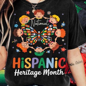 K&ouml;nnte beinhalten: Schwarzes T-Shirt mit einer farbenfrohen Grafik zur Feier des Hispanic Heritage Month. Das Design zeigt einen Schmetterling mit einem lebendigen Fl&uuml;gelmuster, umgeben von Cartoon-Kindern. Der Text enth&auml;lt "National" und "Hispanic Heritage Month."
