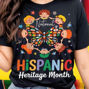 K&ouml;nnte beinhalten: Schwarzes T-Shirt mit einem bunten Schmetterlingsdesign, umgeben von Cartoon-Kindern, die sich an den H&auml;nden halten. Das Shirt zeigt die Worte "National Hispanic Heritage Month" in leuchtenden Farben, die kulturelle Vielfalt feiern.