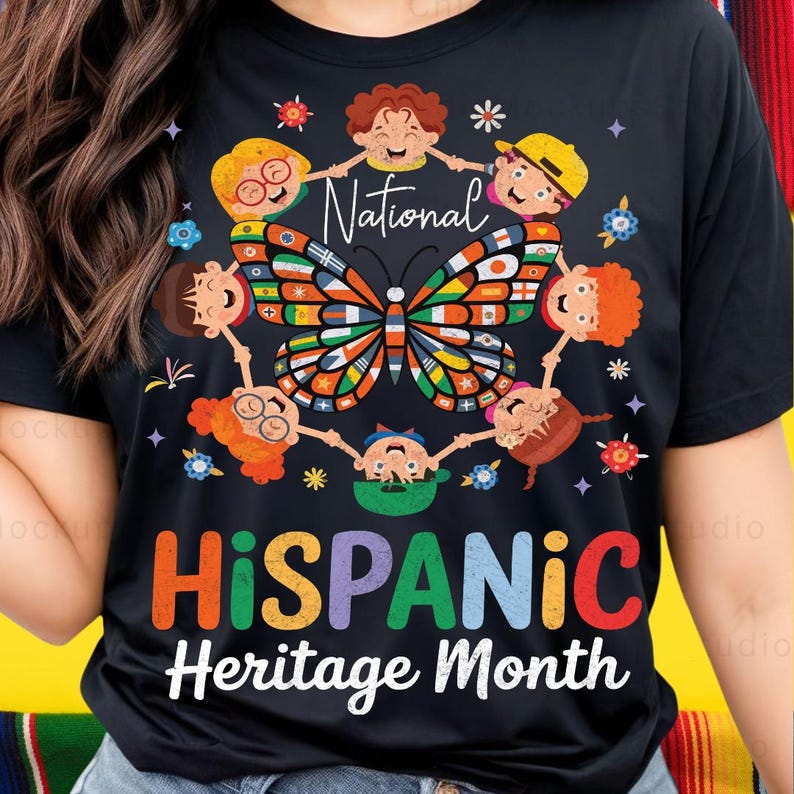 K&ouml;nnte beinhalten: Schwarzes T-Shirt mit einem bunten Schmetterlingsdesign, umgeben von Cartoon-Kindern, die sich an den H&auml;nden halten. Das Shirt zeigt die Worte "National Hispanic Heritage Month" in leuchtenden Farben, die kulturelle Vielfalt feiern.