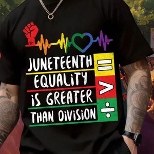May include: A black t-shirt featuring a graphic with a red raised fist, a rainbow heartbeat line, and a heart. The text reads JUNETEENTH EQUALITY IS GREATER THAN DIVISION, accompanied by mathematical symbols for equals, greater than, and division on color blocks.
