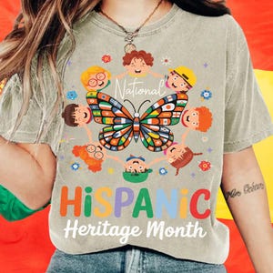 K&ouml;nnte beinhalten: Hellgr&uuml;nes T-Shirt mit einem bunten Schmetterlingsdesign, umgeben von Cartoon-Kindern. Das Shirt tr&auml;gt die Worte "National Hispanic Heritage Month" in einer lebendigen, mehrfarbigen Schrift.