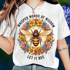 May include: White t-shirt with a colorful floral wreath design surrounding a detailed bee illustration. The text "WHISPER WORDS OF WISDOM" arches above the bee, with "LET IT BEE" below. The bee has yellow and black stripes.