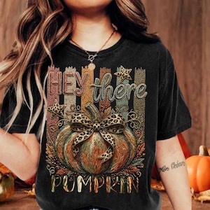 May include: A black t-shirt featuring a graphic print of a pumpkin with a leopard print bow. The design includes vertical stripes in muted autumn colors, two leopard print stars, and the text "HEY there PUMPKIN" in a stylized, distressed font.