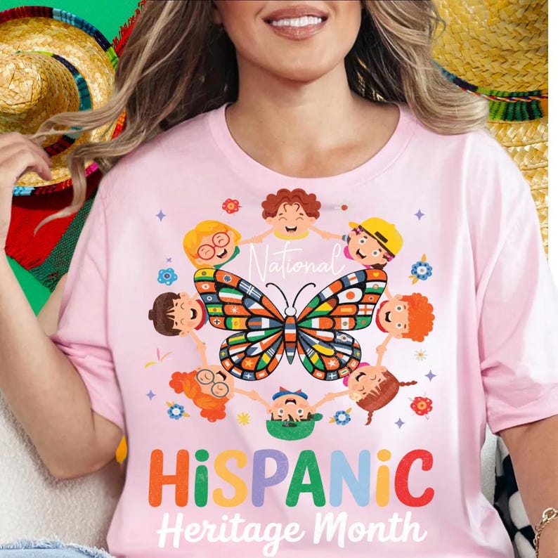 K&ouml;nnte beinhalten: Hellrosa T-Shirt mit einer farbenfrohen Grafik zur Feier des Hispanic Heritage Month. Das Design zeigt einen Schmetterling mit Flaggen, Kindern und den Worten "National Hispanic Heritage Month" in Orange und Wei&szlig;.