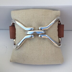 Pode incluir: Uma pulseira de prata com uma tira de couro marrom. A pulseira tem a forma de um oito.