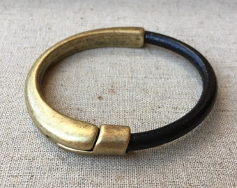 May include: A circular bracelet featuring a smooth, matte gold-toned metal segment and a contrasting dark black band. The metal portion includes a visible hinge mechanism that connects to the black section, creating a continuous, rounded loop design.
