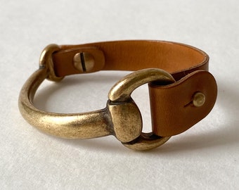 May include: A brown leather bracelet with a brass horse bit buckle. The bracelet is adjustable and has a simple design.