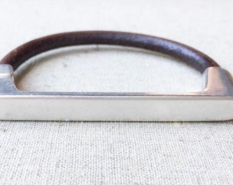 May include: A silver and brown leather bracelet with a flat, rectangular bar design.