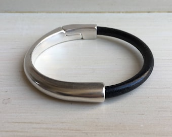 May include: A silver and black leather bracelet with a simple, modern design. The bracelet has a smooth, rounded silver band and a black leather strap.