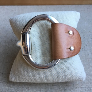 May include: A silver metal bracelet with a leather strap. The leather strap is brown and has two silver rivets. The bracelet is on a white fabric surface.