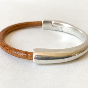 Puede incluir: Un brazalete de cuero marrón con un cierre de metal plateado. El brazalete es simple y elegante.