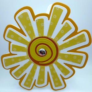 May include: A yellow and orange sun with a white background and a small sparkly jewel in the center. The sun has a spiral design in the center.