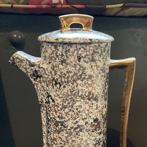 May include: A white ceramic coffee pot with a gold lid and handle. The pot has a speckled pattern of gold and black.