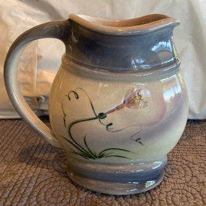 May include: A ceramic pitcher with a light blue glaze and a floral design. The pitcher has a round body and a handle. The floral design features a white flower with green leaves.