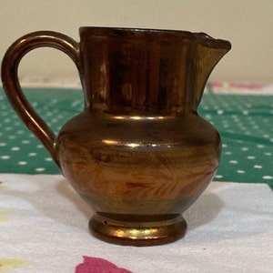 May include: A small, copper-colored ceramic creamer with a handle and a floral design. The creamer is about 4 inches tall and has a rounded body.