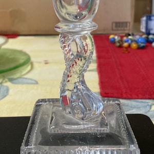 May include: Clear glass candlestick holder with a square base and a decorative, twisted stem. The top portion is a cup shape. The word "Vita" is visible on the top of the candlestick.