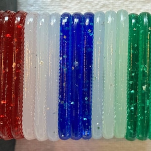 May include: A collection of colorful, glitter-infused bangles. The bangles are arranged in a row, showcasing a vibrant palette of red, white, blue, light green, and dark green. Each bangle has a textured, ridged surface.