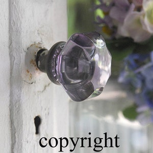 May include: A close-up of a purple glass doorknob with a hexagonal shape. The doorknob is mounted on a white door with a keyhole visible.