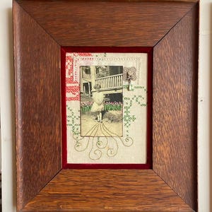 May include: A framed vintage photograph of a child in a garden, with cross-stitch embroidery and red velvet. The wooden frame has a dark brown finish, enhancing the antique aesthetic. The photograph is centered within the frame, creating a nostalgic feel.