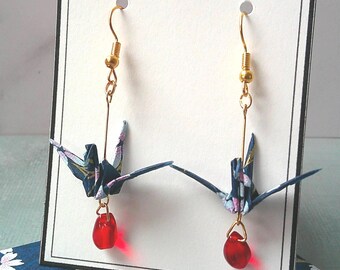 May include: A pair of origami crane earrings. The cranes are navy blue with a floral pattern, suspended from gold-coloured hooks with red teardrop beads. The earrings are displayed on a card with the text "ROSYTOMMY".