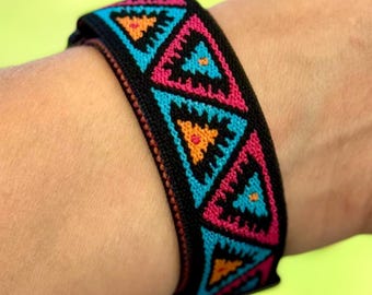May include: A black woven bracelet with a repeating pattern of pink, teal, and orange triangles. The bracelet is worn on a wrist, showcasing the colorful geometric design. The bracelet is approximately 1.5 inches wide.