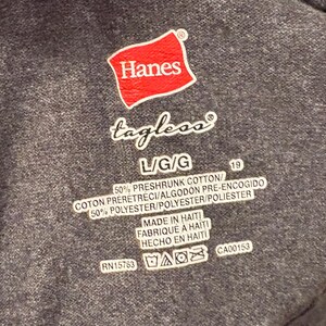 May include: Close-up of a dark grey Hanes tagless t-shirt label. The red Hanes logo is at the top. The label includes size, fabric content, and care instructions. Made in Haiti.