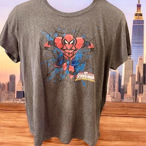 May include: A dark grey t-shirt with a graphic of Spider-Man in a red and black suit, set against a blue web design. The text "SPIDER-MAN WEB-WARRIORS" is printed below the image.