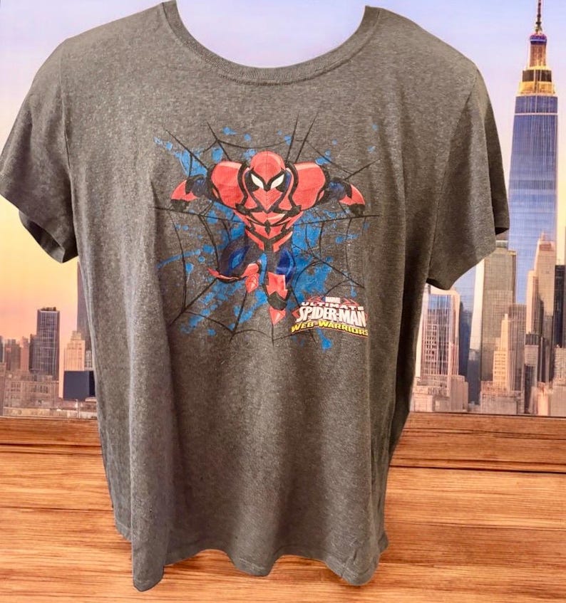 May include: A dark grey t-shirt with a graphic of Spider-Man in a red and black suit, set against a blue web design. The text "SPIDER-MAN WEB-WARRIORS" is printed below the image.