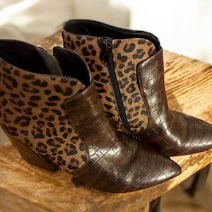 May include: A pair of brown ankle boots with a pointed toe and a textured, crocodile-like pattern. The upper portion of the boots features a leopard print design, and a zipper closure is visible on one boot.